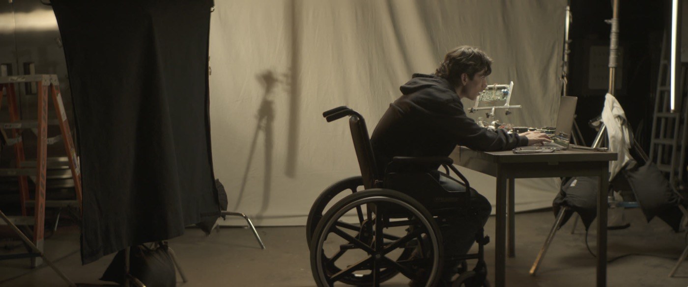 Adam in his wheelchair at a workbench, typing, a circuit board in a clamp beside him